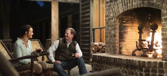 A couple sitting in front of a large, brick outdoor fireplae enjoying wine and whiskey.