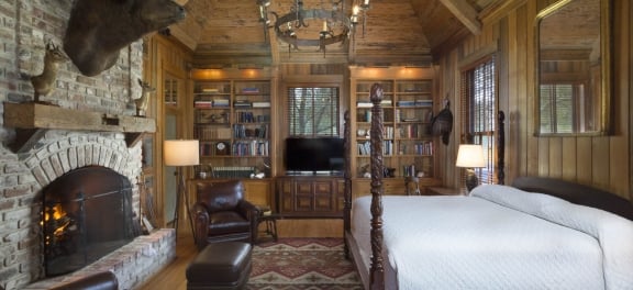 The Broadfield Lodge Master Bedroom, a large wooden four-poster bed in front of a grand brick fireplace. The room is wood panel with vaulted ceilings, mahogany and solid wood furniture. A taxidermy bison head is mounted above the fireplace.