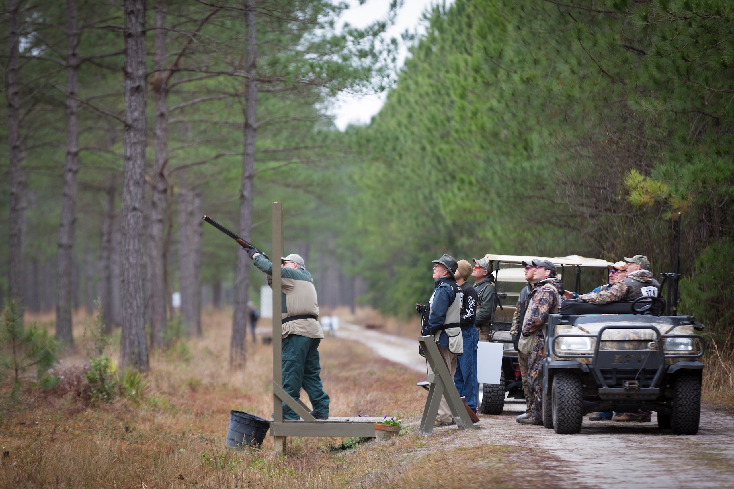 A man shoots a long gun while a small group watches from behind, some sitting on a golf cart.