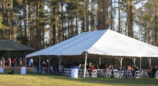 A large outdoor seating area covered with a large white tent at sunset, wooded forest in the background.