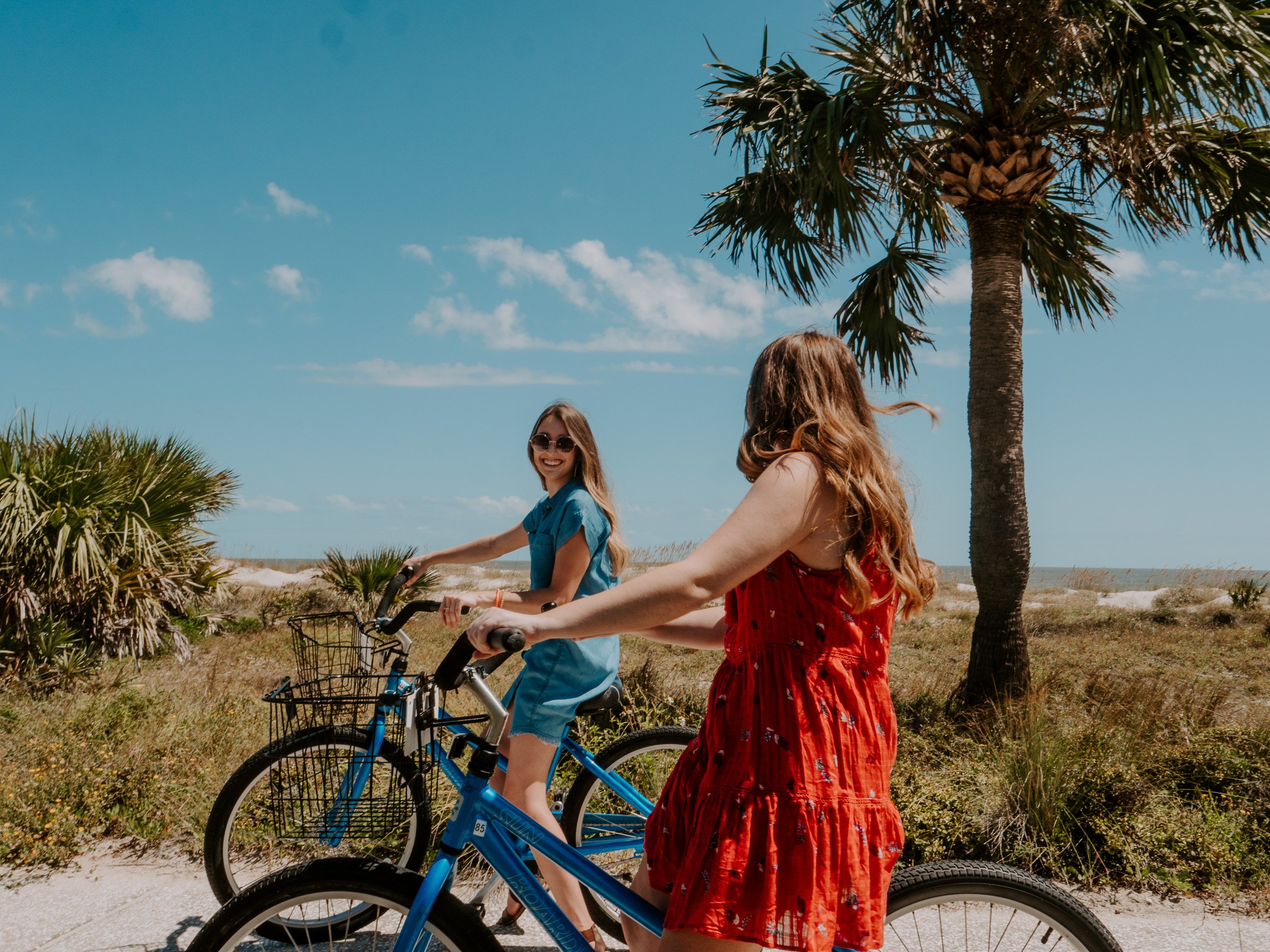 Two women biking along a sandy trail to a beach on a sunny day.