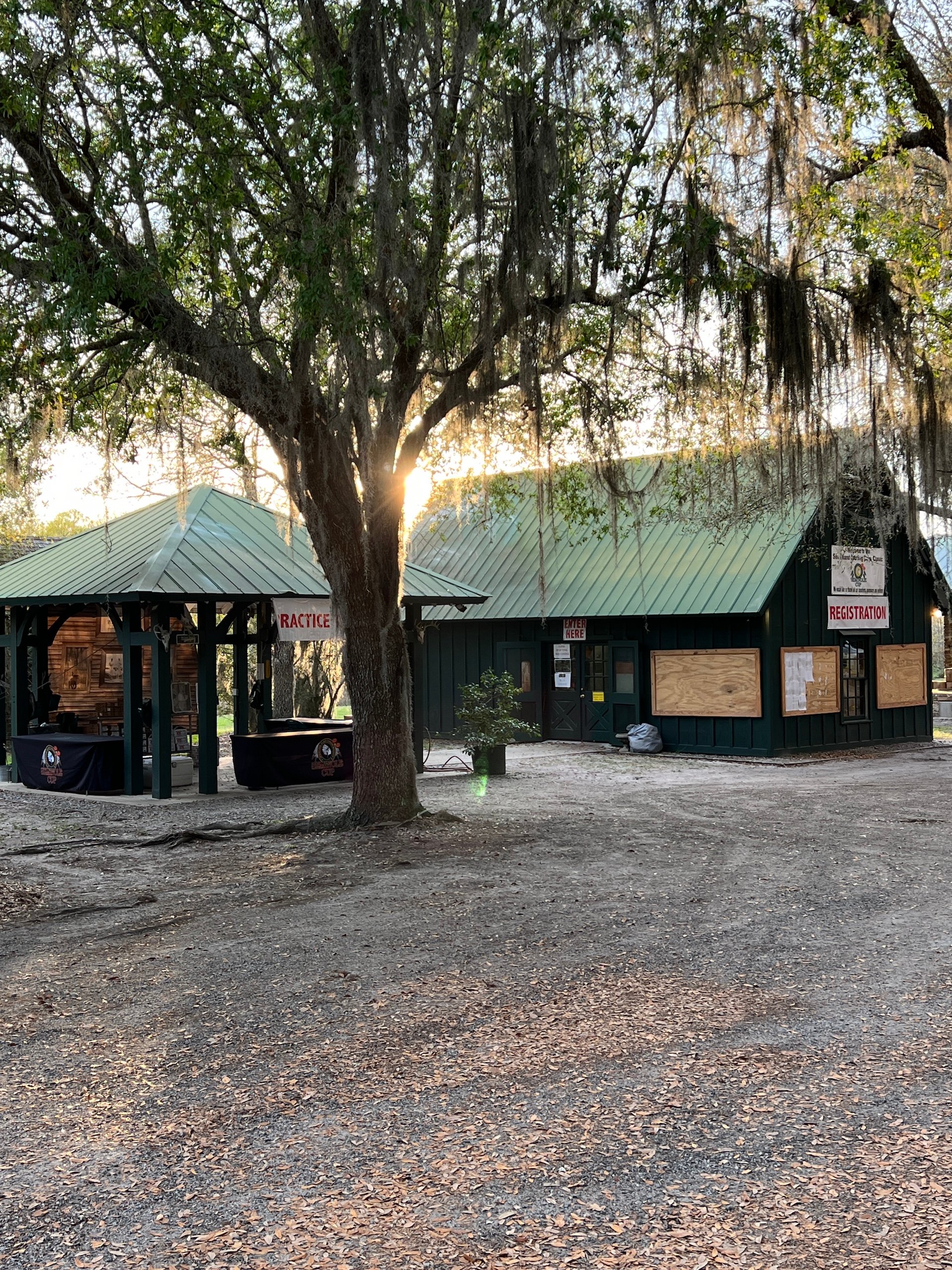 Two small registration buildings on a dirt road behind a willow tree.