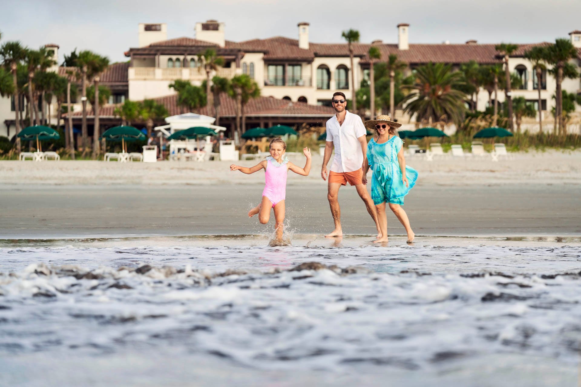 A little girl runs into the water as her parents look on smiling from the beach coastline at Sea Island.