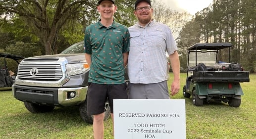 Two men standing behind a reserved parking sign in a field, a pick up truck and golf card behind them.