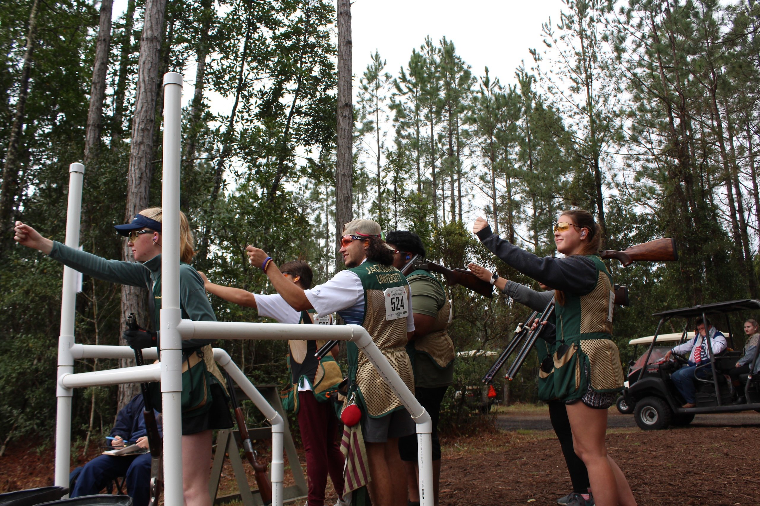 A group of people pointing at a shooting competition in the woods. Spectators watch in the background from golf carts.