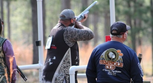 A man fires a long gun during a shooting competition while a judge watches beside him.