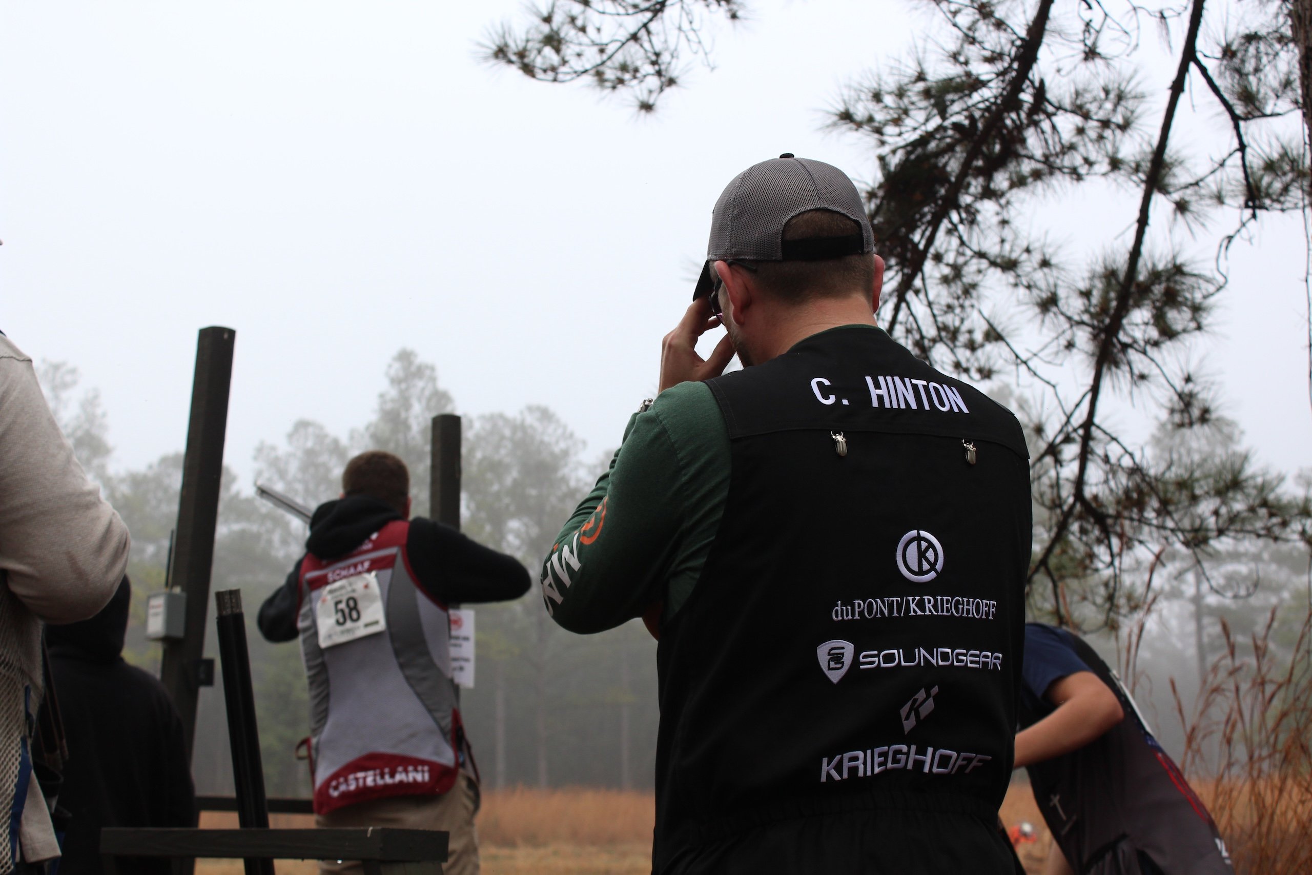 A man covers his ears as he stands behind a competing shooter on a foggy day at Seminole Cup.