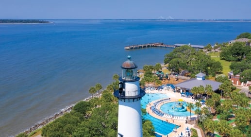 An aerial view of Golden Isles. A tall white lighthouse is seen in the foreground, a pool and splash pad behind it. In the background is blue ocean on a sunny day.