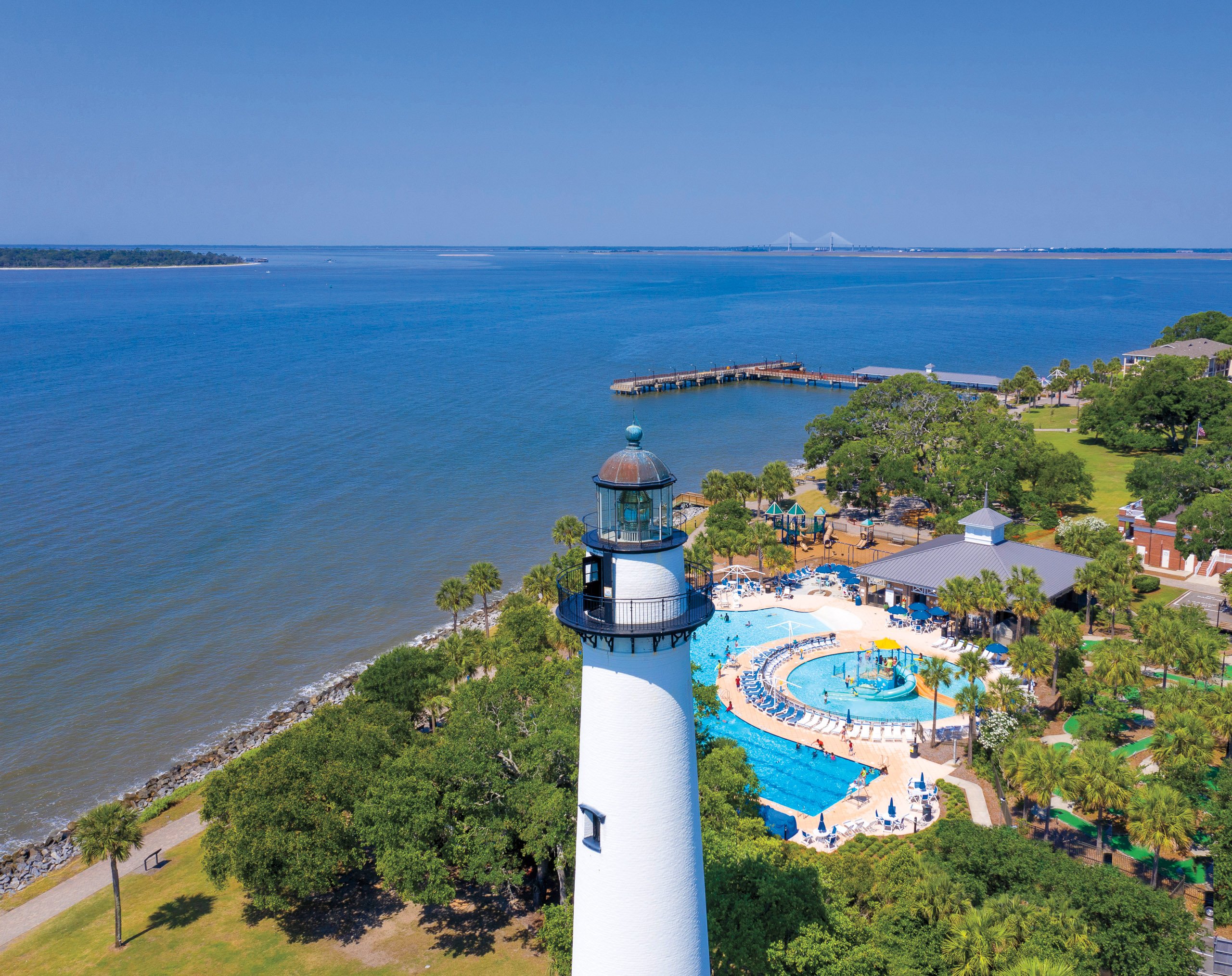 An aerial view of Golden Isles. A tall white lighthouse is seen in the foreground, a pool and splash pad behind it. In the background is blue ocean on a sunny day.