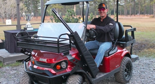 A man sits smiling in the drivers seat of a red EZ-GO golf cart with gun rack attached.