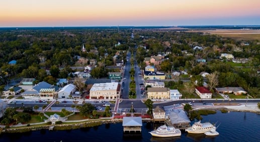 An aerial view of St. Mary's at sunset. Three boats are parked in the water next to the main street. The town sprawls out into more wooded areas.