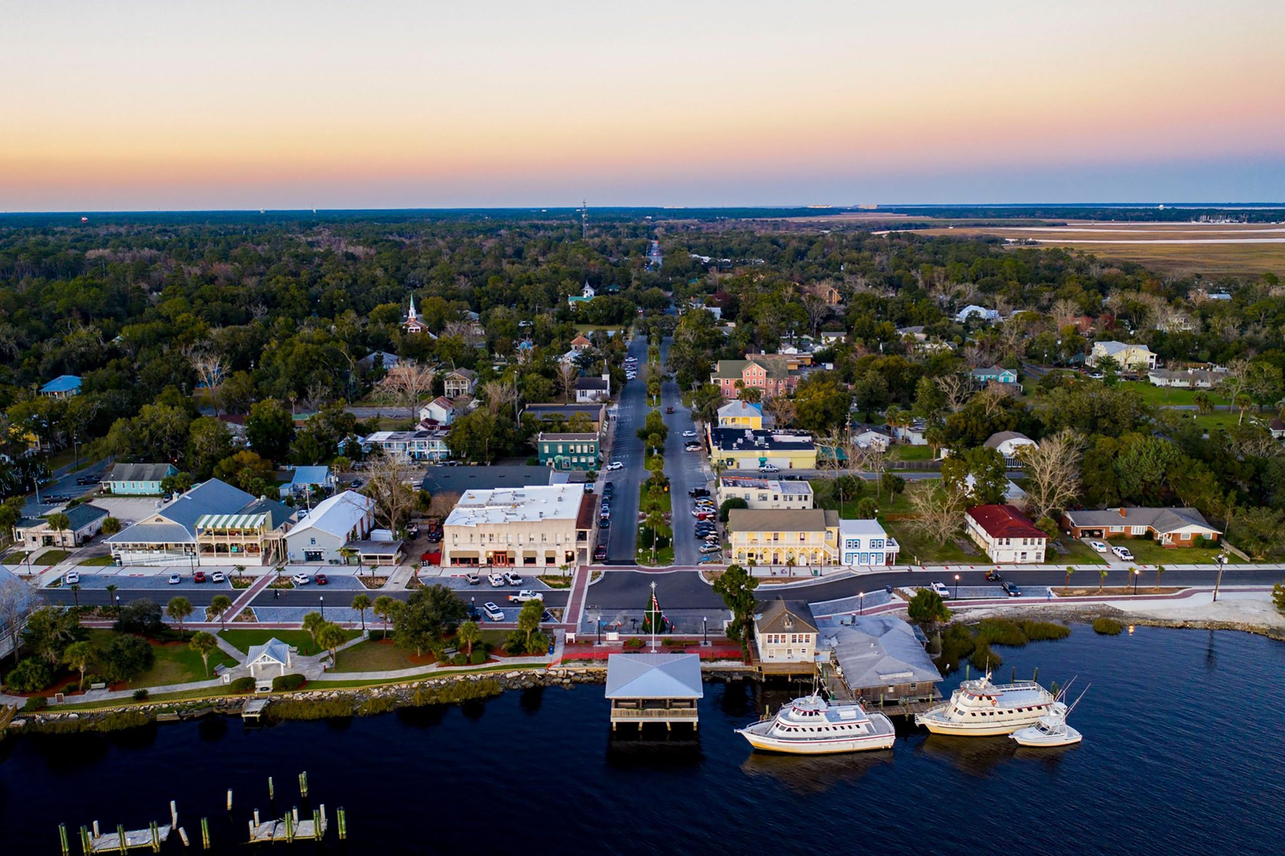 An aerial view of St. Mary's at sunset. Three boats are parked in the water next to the main street. The town sprawls out into more wooded areas.