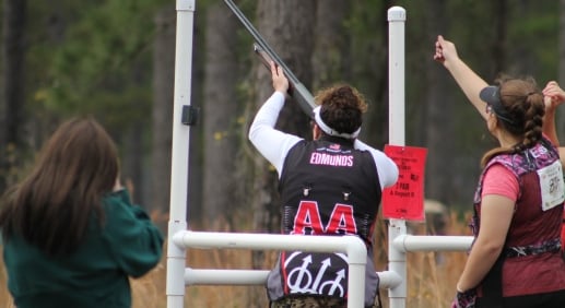 A woman aims a rifle at a competition while others stand behind her watching.